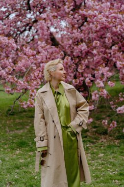 Çiçek açan pembe kiraz çiçeklerinin önünde, yeşil saten elbiseli şık sarışın bir kadın zarafet ve zarafet yayar, baharın ve doğanın güzelliğini büyüleyici bir şekilde yakalar.
