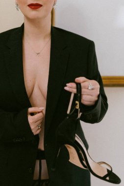 Zarif ceketli ve iç çamaşırlı göz kamaştırıcı bir kadın, yüksek topuklu siyah bir ayakkabı tutuyor. Şık duruşu lüks ve zarafet saçıyor. Şık ve zarafet karışımı bir şey sergiliyor.