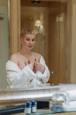 A confident woman with short blonde hair stands in front of a mirror in a white bathrobe, elegantly applying lipstick, reflecting her beauty and poise as part of her daily selfcare routine clipart