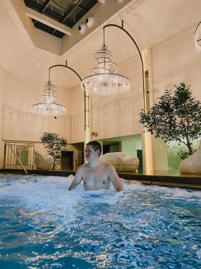 Bir adam lüks bir tatil köyünde, şık dekorlar ve sıcak aydınlatmalarla çevrili kapalı bir yüzme havuzunun rahatlatıcı atmosferini sever. Seyahat, rahatlama ve boş zaman temaları için mükemmel..