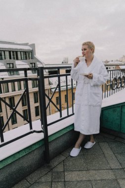 Beyaz bornozlu ve terlikli bir kadın karlı bir çatıda duruyor ve bir fincan kahvenin tadını çıkarıyor. Şehir manzarası arka planda görünür, rahat ve rahat bir tatil sahnesi yaratır.