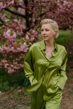 Çiçek açan pembe kiraz çiçeklerinin önünde, yeşil saten elbiseli şık sarışın bir kadın zarafet ve zarafet yayar, baharın ve doğanın güzelliğini büyüleyici bir şekilde yakalar.