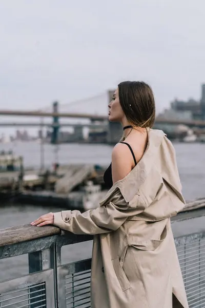 Şık bir trençkot giymiş bir kadın sakin bir şehir ortamında parıldayan suyun kenarındaki uzak bir şehir köprüsüne bakıyor. Trençkot giyen şık bir kadın suyun kenarında durmuş güzel şehir köprüsüne bakıyor.