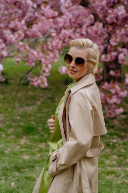 Çiçek açan pembe kiraz çiçeklerinin önünde, yeşil saten elbiseli şık sarışın bir kadın zarafet ve zarafet yayar, baharın ve doğanın güzelliğini büyüleyici bir şekilde yakalar.