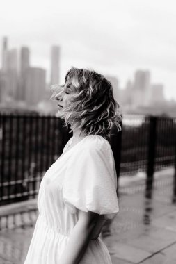 Zarif bir kadın zarif bir şekilde kırsal bir bankta poz veriyor. Yumuşak yağmurun ortasında. Arka planda çarpıcı bir şehir silüeti var.