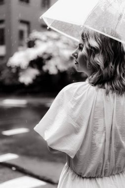 Zarif beyaz bir şemsiye tutan sakin bir kadın yağmurlu bir şehir arka planında derin düşüncelere dalmış gibi görünüyor.