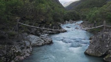İskandinavya 'da güzel bir insansız hava aracı uçuşu, hızlı bir dağ nehri, mavi köpüklü suda büyük kayalar. Norveç 'te ahşap asma köprüsünde yürüyen maceracı, genç yürüyüşçü kızın görüntüsü.