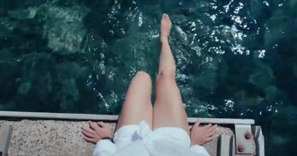 女人的头像坐在码头上 在水里摇晃着脚 暑假让人兴奋 节日气氛的灵感 美丽的绿松石海水 游客在水里活动腿以感到清爽 — 图库视频影像