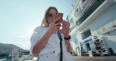 Geniş açılı çekimde genç bir kadın tatil sırasında GPS 'e bakıyor. Güneşli bir yaz günü, kumsalda beyaz gömlekli bir kadın telefonda İspanyol tatil belgesi, sosyal medya ya da harita kullanarak yolunu bulur.. 