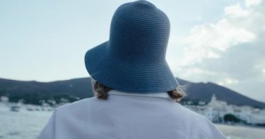 Otantik ve gerçek kadın turist portresi. Bohem beyaz kıyafeti ve mavi kova şapkalı. Kameraya bak, gülümse, Akdeniz kıyısındaki tatil köyünde manzaranın tadını çıkar.