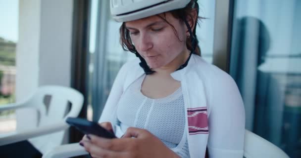 サイクリングギアスポーツウェアの若い女性は カフェや自宅で座っています Gpsコンピュータから彼女のスマートフォン上の乗り心地やルートをアップロードします フィットネスの進歩を追跡するためにプロの女性サイクリングアマチュア使用技術 — ストック動画