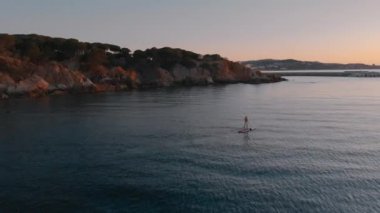 Aerial drone shot of athlete training in the sea on stand up paddle board in sunrise or sunset. Recreational adventure for active man in beautiful mountain landscape. 