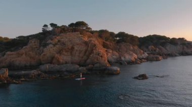 Sunrise or sunset aerial drone shot of man paddle board near coast in Europe. Cinematic mountains view. Athlete on stand up SUP board in blue sea explore European shores