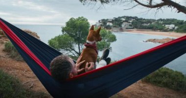 Ağaçlar ve ormanlarla çevrili bir tepede, hamakta sallanan Basenji köpeği olan bir adamın arkası. Mavi denizin huzurlu manzarasından hoşlanır, yaz güneşinde meditasyon yapar ve dinlenir.. 