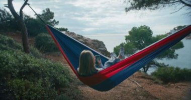 Kadın hamakta telefonla sallanıyor, ormanın yemyeşil yeşillikleriyle çevrili. Tepede, deniz manzaralı bir yerde bir dinlenme ve meditasyon anı buluyor. Beslemesini mesaj atıyor ve kaydırıyor..