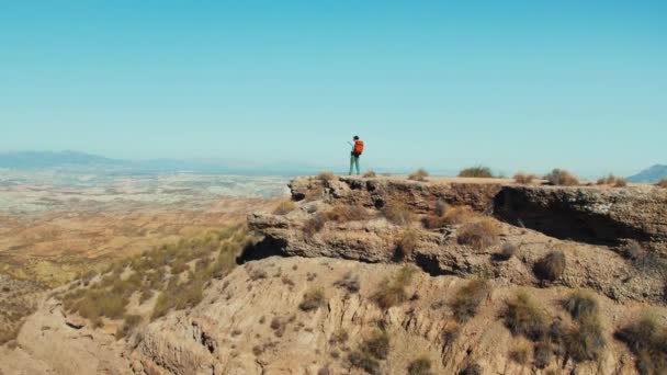 美しい景色に囲まれた男性冒険家は 砂漠の丘の上に立っています 彼の頂上へのハイキングは 遠くの山の平和的な景色のためにそれの価値があった 空中射撃 — ストック動画