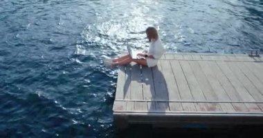 Kadın dizüstü bilgisayarın yanındaki iskelede rahatça oturur, denize bakar, sakin bir yaz gününün ve uzak bir işin tadını çıkarır. Huzurlu mutluluk, tatmin ve özgürlük anı.