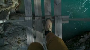 Yürüyüş botları giymiş bir adam vahşi dağ nehri üzerindeki ahşap asma köprünün üzerinden geçiyor. Destansı yürüyüş macerası ya da açık hava keşfi konsepti. Ayakkabılar ahşap köprüde yürür