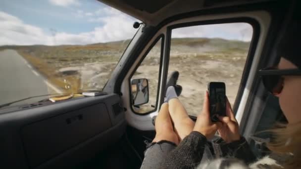 車やキャンパーバンの助手席の若い女性旅行者は窓から足をぶら下げてスマートフォンで写真を撮る 北欧諸国のロードトリップ夏の冒険 — ストック動画