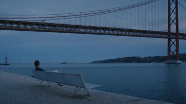İnsanoğlunun kopyası ile güzel bir sinematik çekim nehir kıyısındaki bankta oturuyor, Lizbon 'daki 25 Nisan tarihli metal köprünün üzerinden bakıyor. Huysuz gün batımı, yoğun şehir hayatında rahatlatıcı bir an.