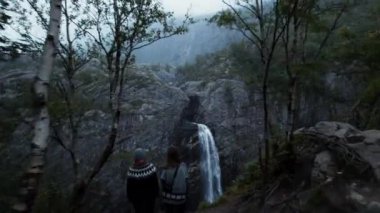Uzun bir yürüyüşten sonra Norveç ulusal parkındaki destansı şelaleye bakan iki turist uçurumun kenarında duruyor. Huysuz sonbahar ormanında şelalenin insansız hava aracı görüntüsü.