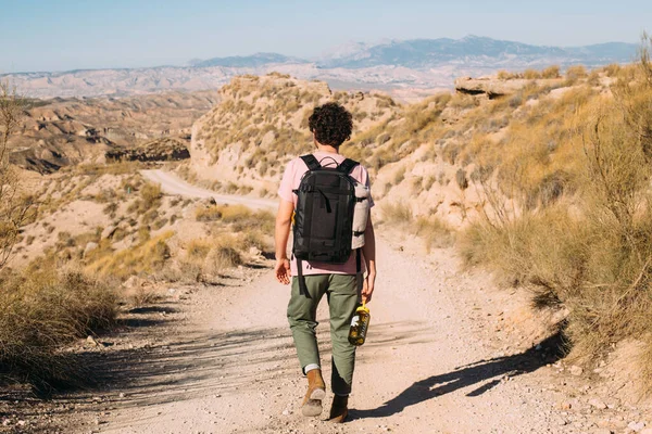 若い男が離れてカメラから 革のブーツで 黒のバックパックを着用し 空のほこりの多い砂漠の道に水のフラスコを運ぶ歩く ムーディ アドベンチャー ハイキングのための暑い晴れた日 — ストック写真
