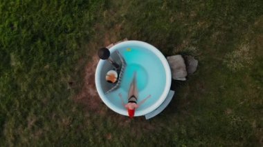 Isıtılmış ahşap jakuzide bikinili ve bereli genç bir kadının kameraya baktığı uzaktan kumandalı görüntüyü büyüt. Doğayla çevrili köylü inzivası. Glamping deneyimi, kaplıca kaçışı.