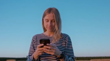 Güzel genç bir kadın gün batımında telefonu kullanır. Mavi gökyüzü arka planında akıllı telefondan mesaj atan doğal görünümlü bir kız. Genç gülümsedi ve internette gezinirken mutlu oldu.