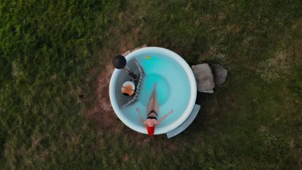 放大无人驾驶飞机拍摄的年轻女子抬头看着相机 穿着比基尼坐在加热的木制热浴缸里 被大自然包围的乡村撤退 有打盹的经验 春温泉逃脱 — 图库视频影像