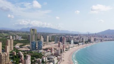 Yüksek katlı turistik konaklama binaları olan Akdeniz kıyısındaki turist kasabasının yavaş çekim yapan insansız hava aracı görüntüleri. Güneşli sahil kıyısında tatil kiraları