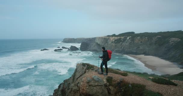 赤いバックパックとトレッキングポールで男性のハイカーのショットを確立する広い角度は 海の風景を見下ろす崖の端に立っています 壮大な西海岸のトレイルで一人でハイキングする男 — ストック動画
