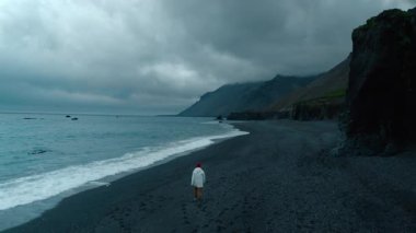 Beyaz ceketli ve kırmızı bereli bir adamın, İzlanda 'daki siyah kumsalda yürüdüğü karamsar, karamsar, siyah bir hava görüntüsü. Gri gökyüzü ve güçlü rüzgar, yaklaşan fırtına konsepti