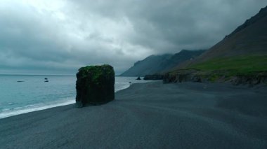 İnsansız hava aracı, İzlanda 'nın ünlü turistik merkezi Laekjavik sahilinde tek başına destansı bir kaya parçasıyla uçuyor. Yağmurlu bulutlar ve gökyüzünde martılarla karamsar ve sinematik gri bir gün.