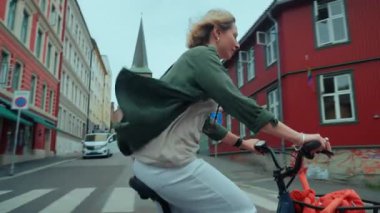 Neşeli gülümseyen ve gülen genç kadın, renkli evleri olan elektrikli bisikletiyle şehir sokaklarında dolanıyor. Çevrede dolaşan bisikletli bir banliyö bisikletçisinin yan fotoğrafı.