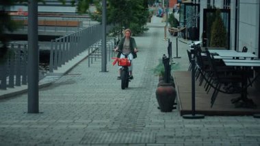 Küçük elektrikli kargo bisikletiyle kameraya doğru giden sevimli genç bir kadın. Yaz boyunca şehirde gidip gelme ve bisiklet sürme