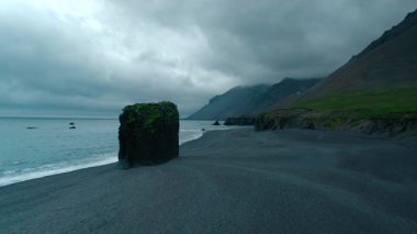 İnsansız hava aracı, İzlanda 'nın ünlü turistik merkezi Laekjavik sahilinde tek başına destansı bir kaya parçasıyla uçuyor. Yağmurlu bulutlar ve gökyüzünde martılarla karamsar ve sinematik gri bir gün.