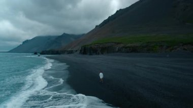Beyaz önlüklü, kırmızı şapkalı, kasvetli bir günde karanlık volkanik kumsalda yürüyen yalnız üzgün bir adam. İzlanda manzarası, sert ve sert iklim koşulları