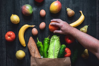 Ahşap bir arka planda sebze, meyve, yumurta ve ekmek dolu bir alışveriş çantasından meyve toplayan bir el. Sağlıklı yemek seçenekleri. Çevrimiçi süpermarket siparişi. Koyu yiyecek fotoğrafçılığı.