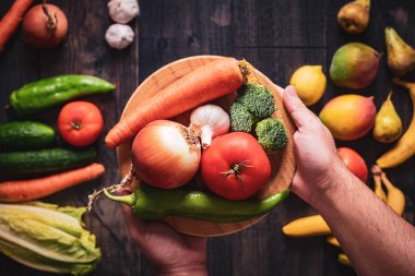 Ahşap bir arka planda meyve ve sebzelerle dolu bir tabak dolusu sebzeyi tutan ellerin yakın çekimi. Taze ve doğal yiyecekler için sağlıklı beslenme ve aşk