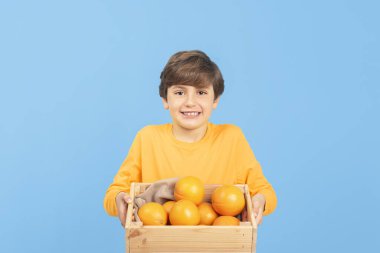 Sarı süveter giymiş, mavi arka planda izole edilmiş portakal dolu bir kutu tutan yakışıklı çocuk. Sağlıklı meyve tüketimi