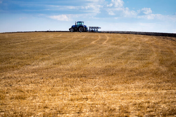 A modern blue tractor with an attached reversible plow with a husking roller is plowing a field on which the spring grain crop has just been harvested. Midsummer in central Ukraine.