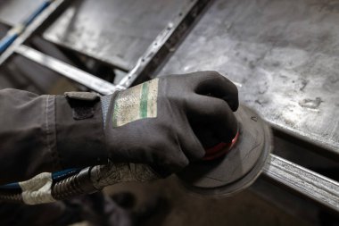 Bir işçinin eldivenli eli, atölyedeki bir tezgahtaki metal çerçevenin eklemlerini pürüzsüz hale getirmek için bir zımpara makinesi kullanır..