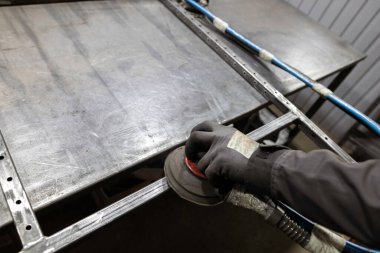 Bir işçinin eldivenli eli, atölyedeki bir tezgahtaki metal çerçevenin eklemlerini pürüzsüz hale getirmek için bir zımpara makinesi kullanır..