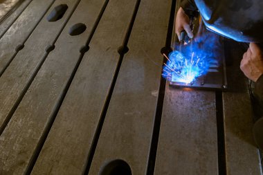 Bir kaynakçı, otomatik kaynak başını tutuyor, ve bir asistan, metal parçaları sıkıca kenetliyor, atölyedeki parlak kıvılcımlarla otomatik kaynak işlemini bir çalışma tezgahında gerçekleştiriyor..