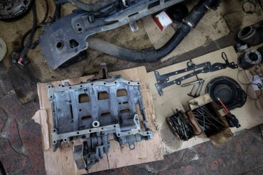 Parçalanmış bir dizel kamyon motoru işlek bir tamirhanede ya da depoda ahşap desteklerin üzerinde yatıyor ve arka planda eğimli bir kamyon taksisi görünüyor.