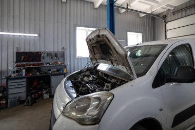 Parçalanmış bir dizel kamyon motoru işlek bir tamirhanede ya da depoda ahşap desteklerin üzerinde yatıyor ve arka planda eğimli bir kamyon taksisi görünüyor.