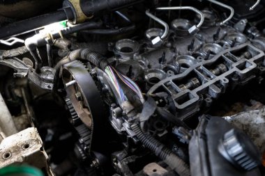 Parçalanmış bir dizel kamyon motoru işlek bir tamirhanede ya da depoda ahşap desteklerin üzerinde yatıyor ve arka planda eğimli bir kamyon taksisi görünüyor.