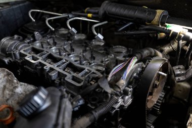 Parçalanmış bir dizel kamyon motoru işlek bir tamirhanede ya da depoda ahşap desteklerin üzerinde yatıyor ve arka planda eğimli bir kamyon taksisi görünüyor.