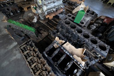 Parçalanmış bir dizel kamyon motoru işlek bir tamirhanede ya da depoda ahşap desteklerin üzerinde yatıyor ve arka planda eğimli bir kamyon taksisi görünüyor.