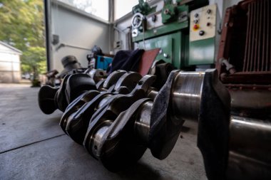 Parçalanmış bir dizel kamyon motoru işlek bir tamirhanede ya da depoda ahşap desteklerin üzerinde yatıyor ve arka planda eğimli bir kamyon taksisi görünüyor.